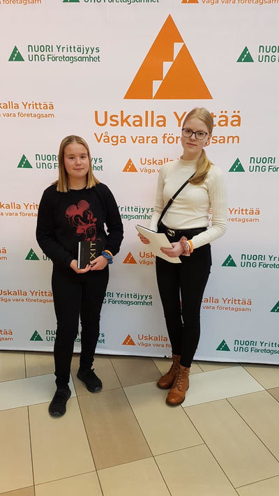 Emilia Viikonkoski ja Jessi Aalto seisovat palkintojensa kanssa mainosseinän edessä Uskalla yrittää -tapahtumassa.