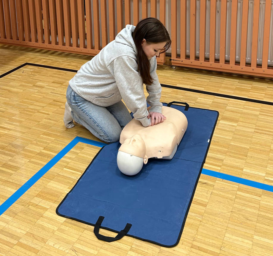 Harmaahupparinen, pitkähiuksinen tyttö antaa elvytystä nukelle sinisen alustan päällä.