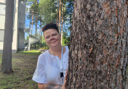 Nainen kurkistaa männynrungon takaa. Naisella on valkoinen pusero, tummat, lyhyet hiukset ja iso tatuointi käsivarressa.