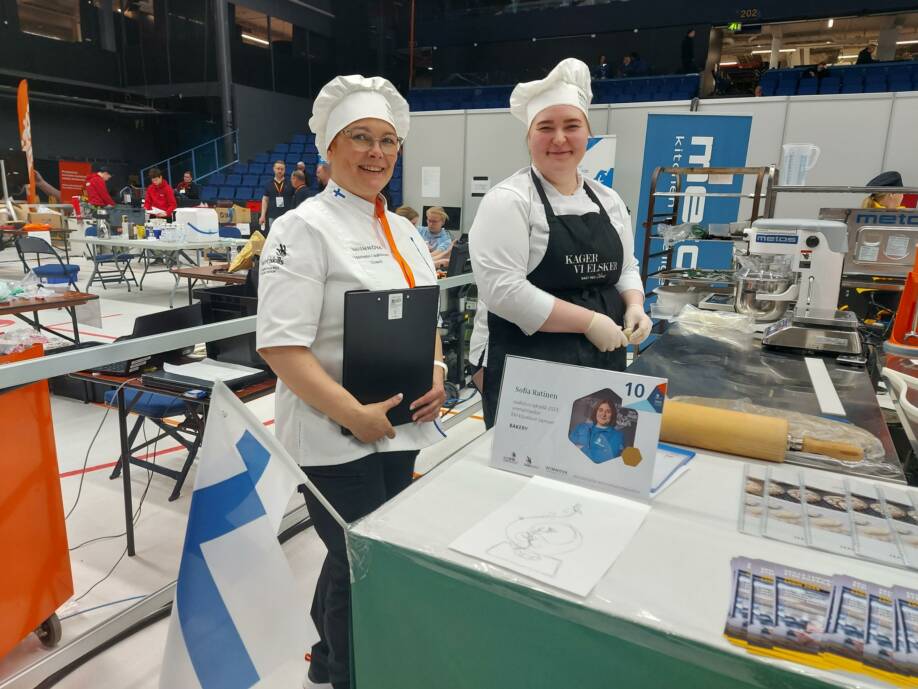 Kaksi naista leipuri-kondiittorien työasussa kisahallissa Taitaja2023-tapahtumassa. Etualalla Suomen lippu.