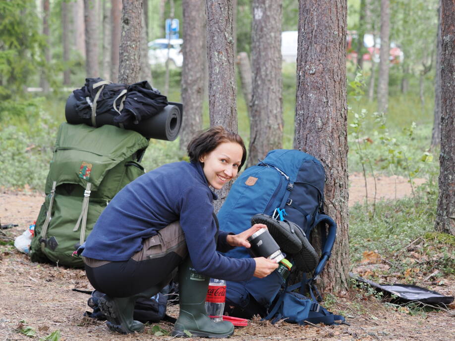 Nuori nainen, jolla on tummanruskea, polkkapituinen tukka kyykyssä sinisen rinkkansa vieressä metsässä. Vieressä on metsänvihreä rinkka, jossa on kiinni vaihtovaatteita ja patja rullalla.