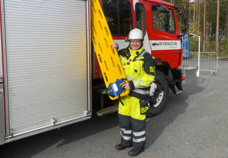 Hymyilevä nainen seisoo paloauton vieressä päällään huomiovärinen työasu ja päässä valkoinen kypärä. Hänellä on keltainen pelastusväline kädessä. Paloauton etuoven kohdalla on WinNovan logotarra.