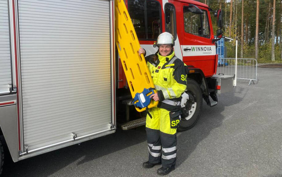 Hymyilevä nainen seisoo paloauton vieressä päällään huomiovärinen työasu ja päässä valkoinen kypärä. Hänellä on keltainen pelastusväline kädessä. Paloauton etuoven kohdalla on WinNovan logotarra.