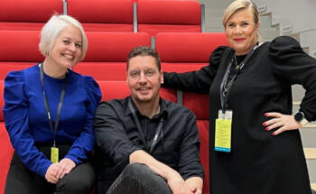 Kuvassa kolme ihmistä auditorion punaisten tuolien edessä. Vasemmalla vaaleahiuksinen nainen sähkönsinisessa puserossa, keskellä silmälasipäinen mies mustassa paidassa ja oikealla seisomassa nainen mustassa tunikassa.